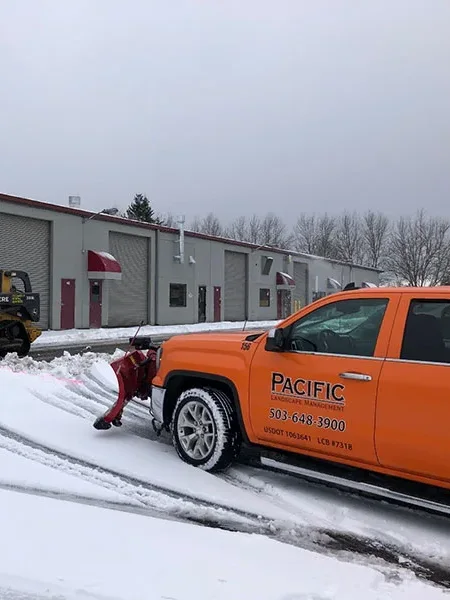 A utility tractor and an orange pickup truck with a snowplow clear snow in front of a row of industrial buildings with red awnings. The truck has branding for Pacific Landscape Management.