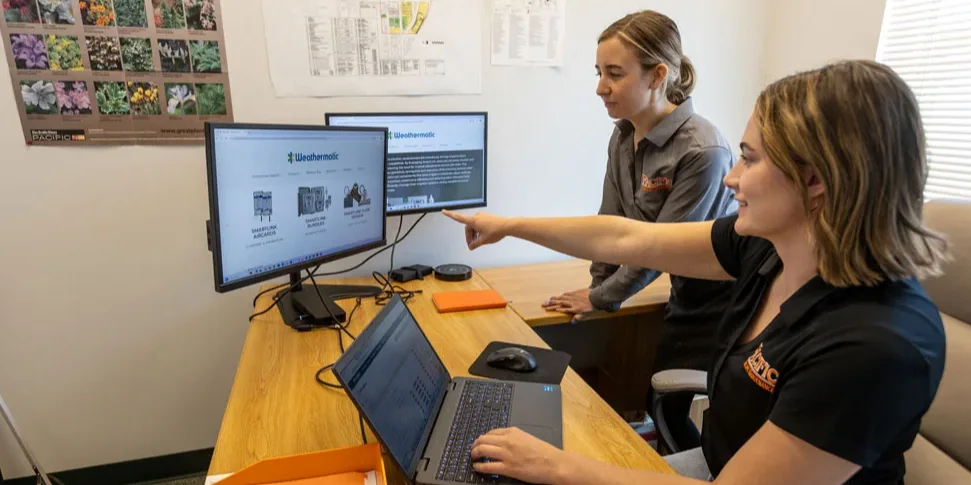 Two women working together at a desk, one pointing at a dual-monitor setup displaying technical information, while the other types on a laptop. Posters and plans are on the wall.