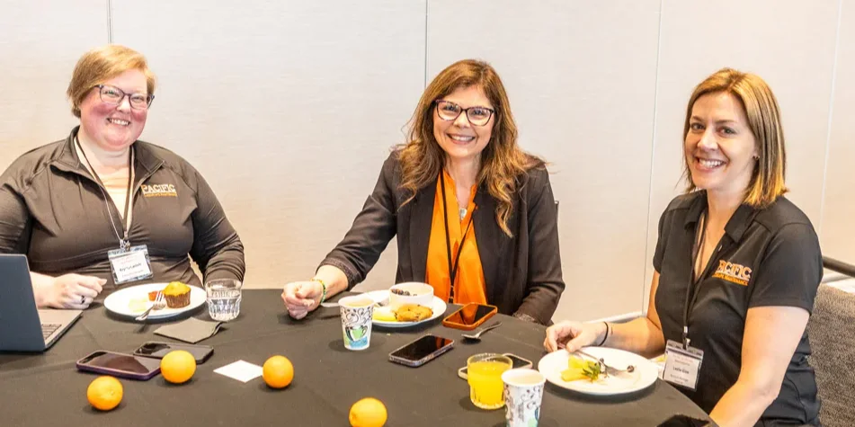 Three women sitting at a round table with food, drinks, and oranges, smiling at the camera. Two wear black shirts with name tags; the other, center, wears a blazer with an orange top.