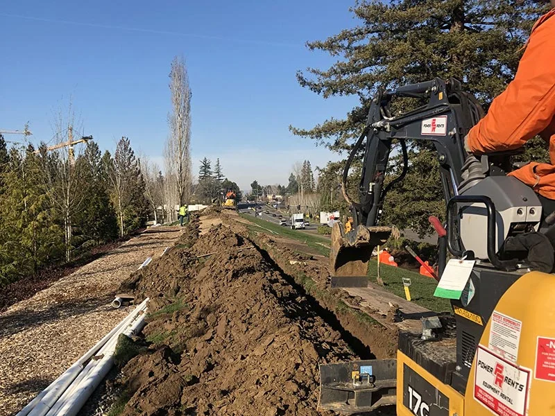 Construction worker operating a small excavator to dig a trench along a road. The trench is filled with dirt, with construction materials and trees lining the pathway on a sunny day.