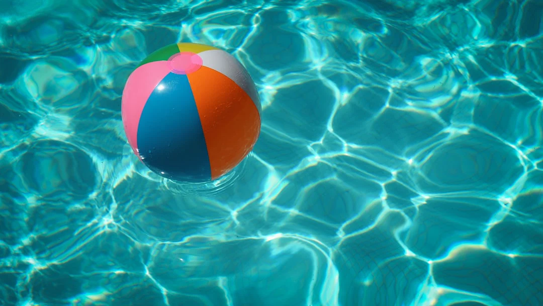 A colorful beach ball floats on clear blue water, with sunlit ripples creating patterns on the pool's surface.