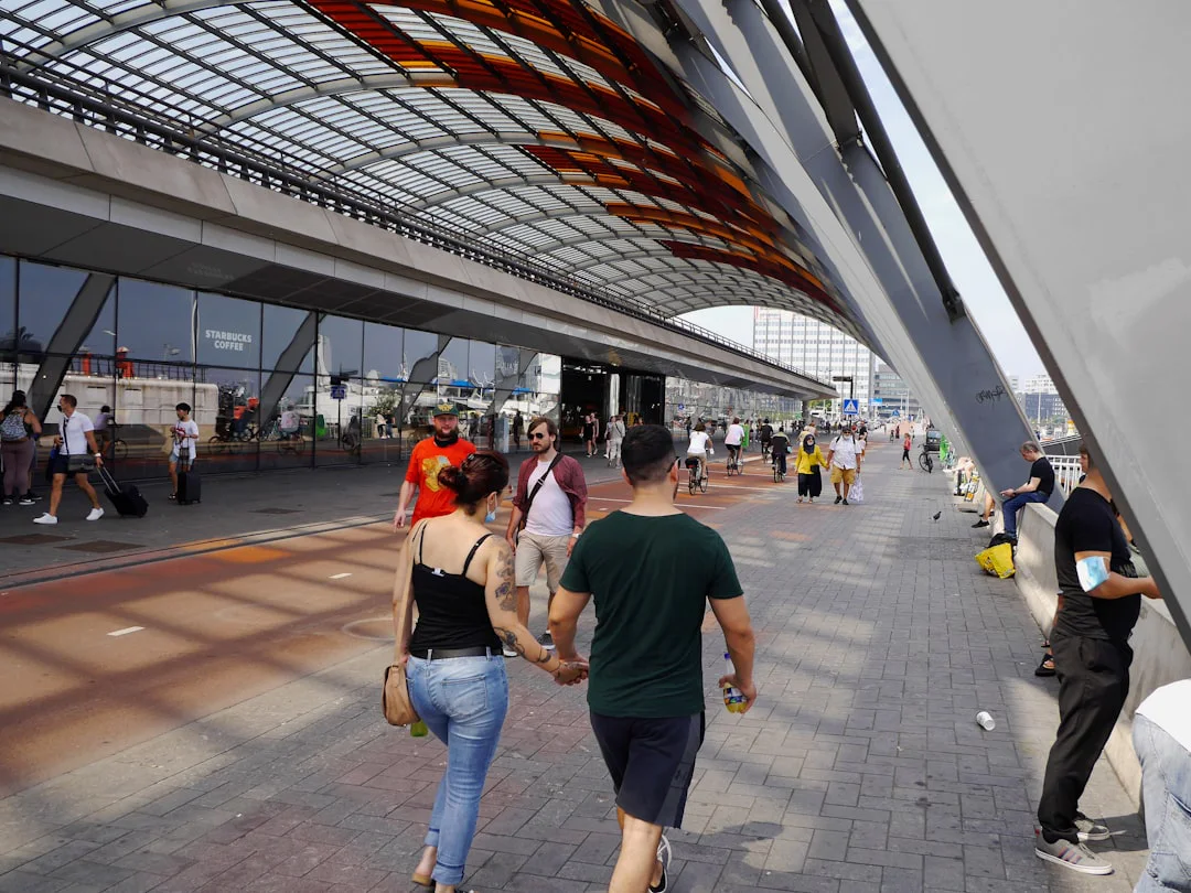 People walk and cycle along a busy, modern bridge with a curved, orange-tinted glass roof. A Starbucks is visible in the background, adding to the urban atmosphere.