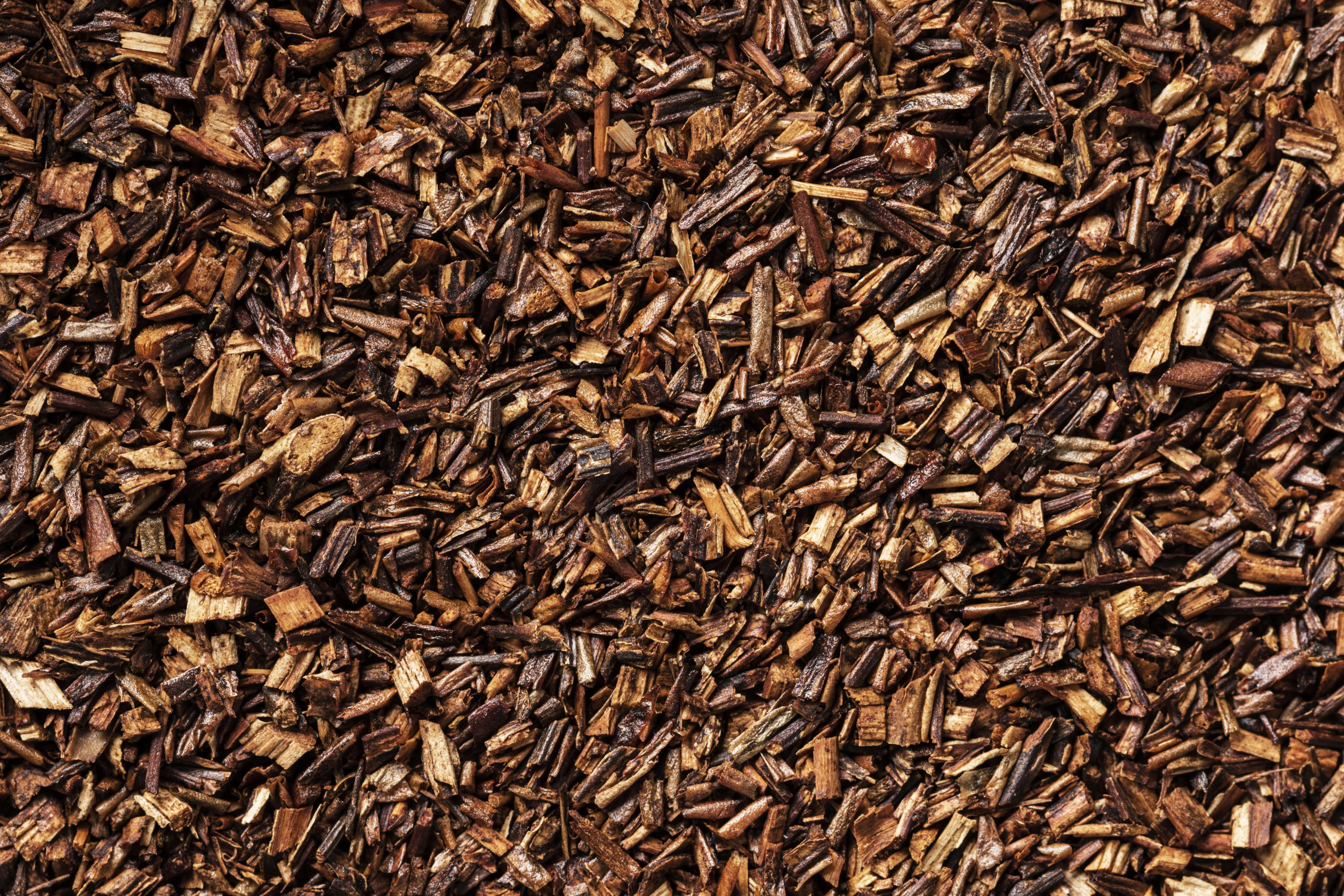 Close-up of scattered rooibos tea leaves, showcasing a mix of small, irregularly shaped, dark brown pieces with a textured and dry appearance.