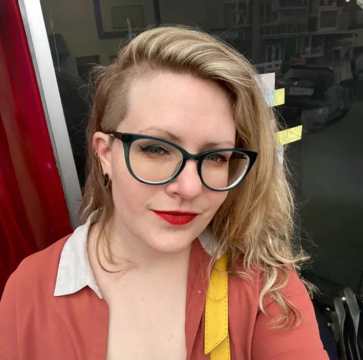 A person with long blonde hair and an undercut, wearing glasses and a red lipstick, is standing outside a building, partially visible through a window. The person has a casual expression.