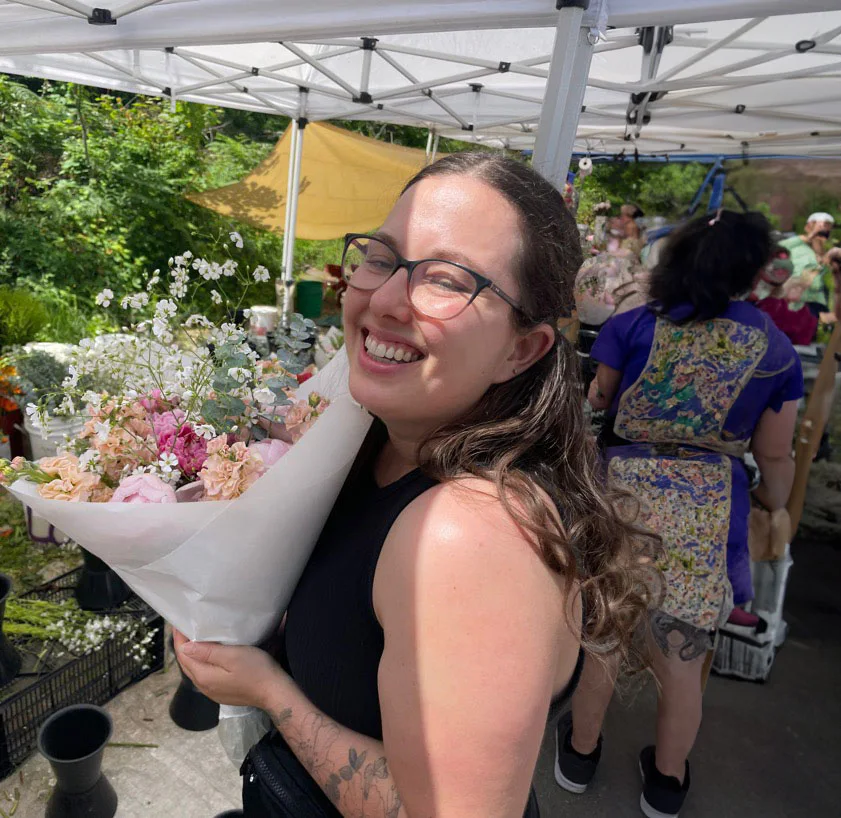 A young woman with glasses and long brown hair smiles while holding a bouquet of flowers under a white canopy. Other people are visible in the background, with greenery around.