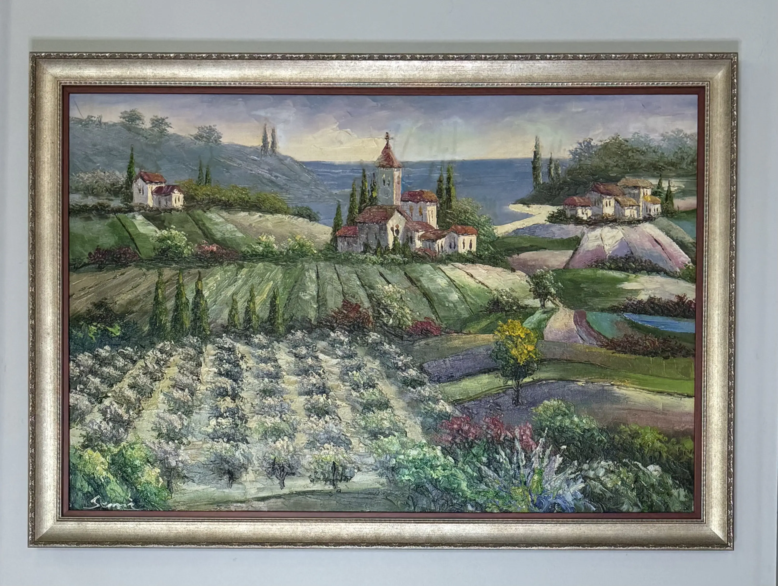 Framed landscape painting depicting rolling hills and fields with trees and small cottages under a cloudy sky, featuring a prominent tree in the foreground.