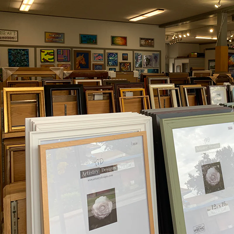 A store with rows of picture frames in various colors and sizes on display. Framed artwork is visible on the back wall, and the ceiling has fluorescent lighting.