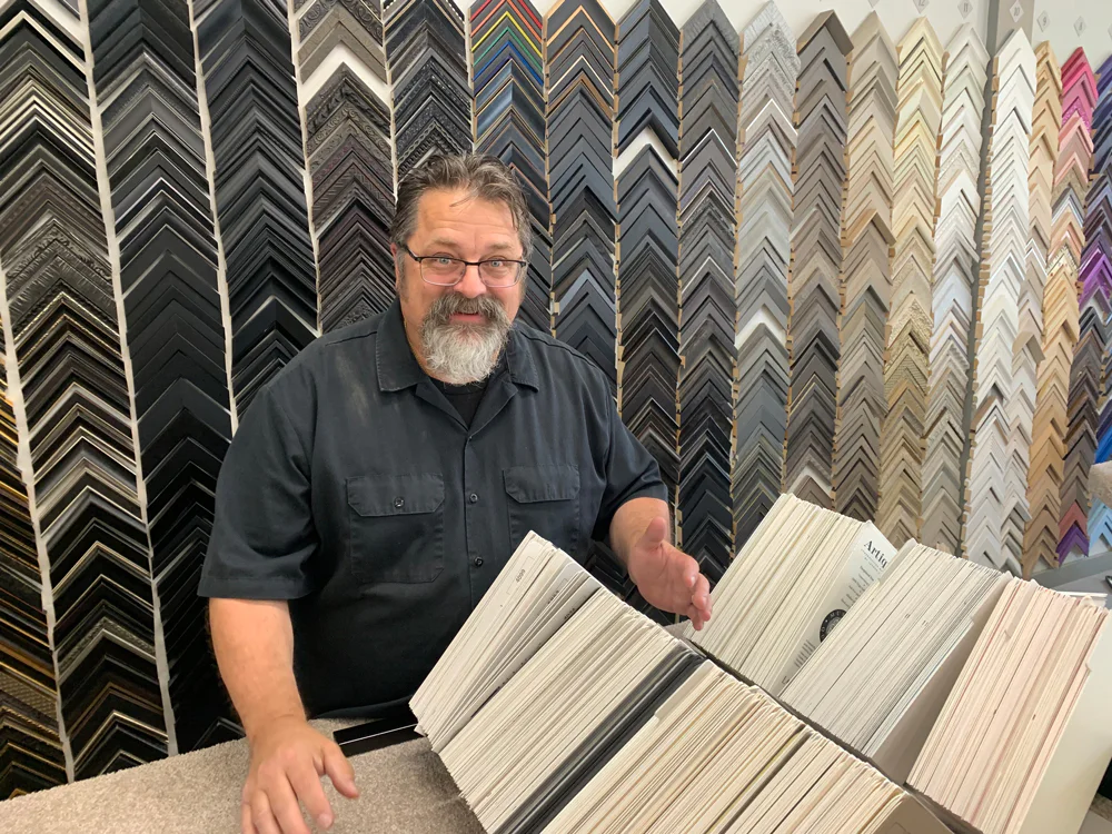 A person with glasses and a beard stands behind a counter displaying various picture frame corners. He is holding an open frame catalog, and there is a wall of frame samples in the background.