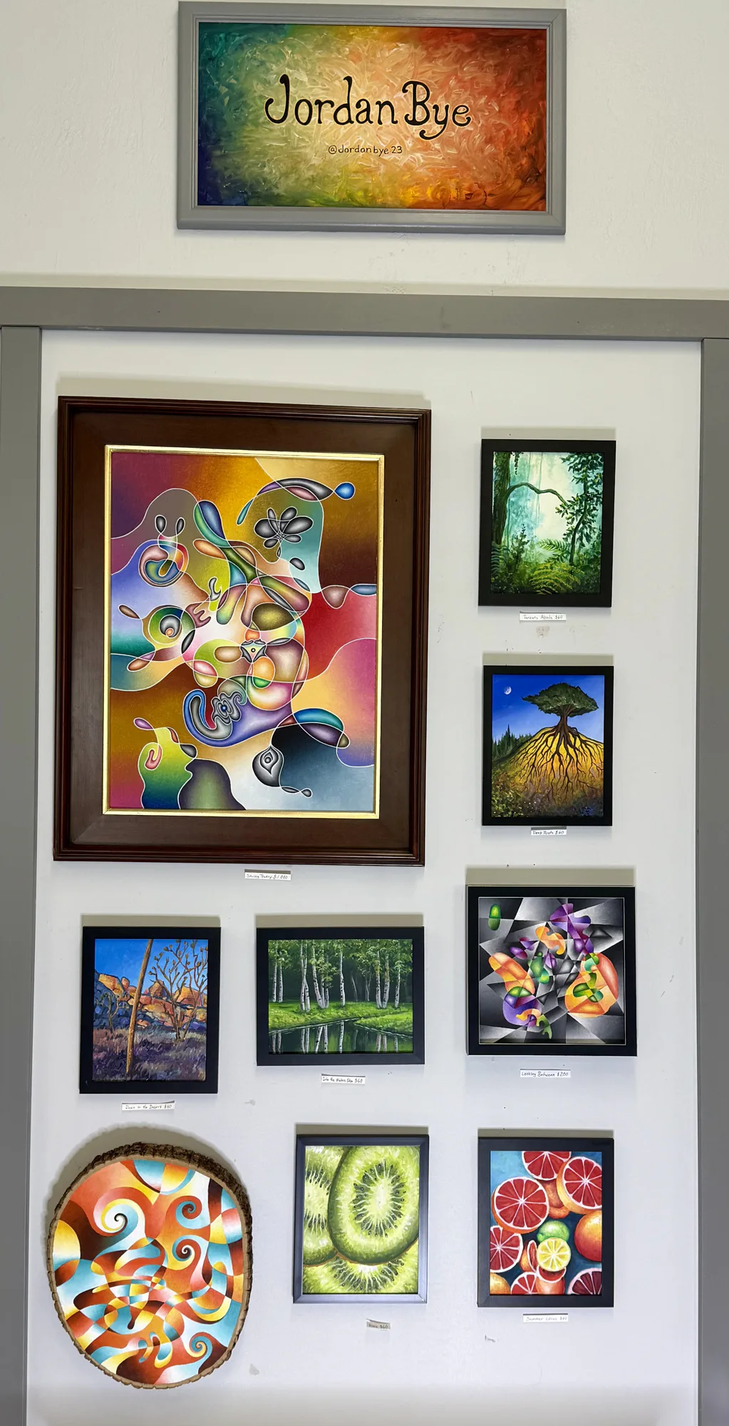 Wall display of colorful abstract, landscape, and fruit paintings by Jordan Bye. The central piece features swirling shapes, surrounded by smaller frames with diverse nature themes.