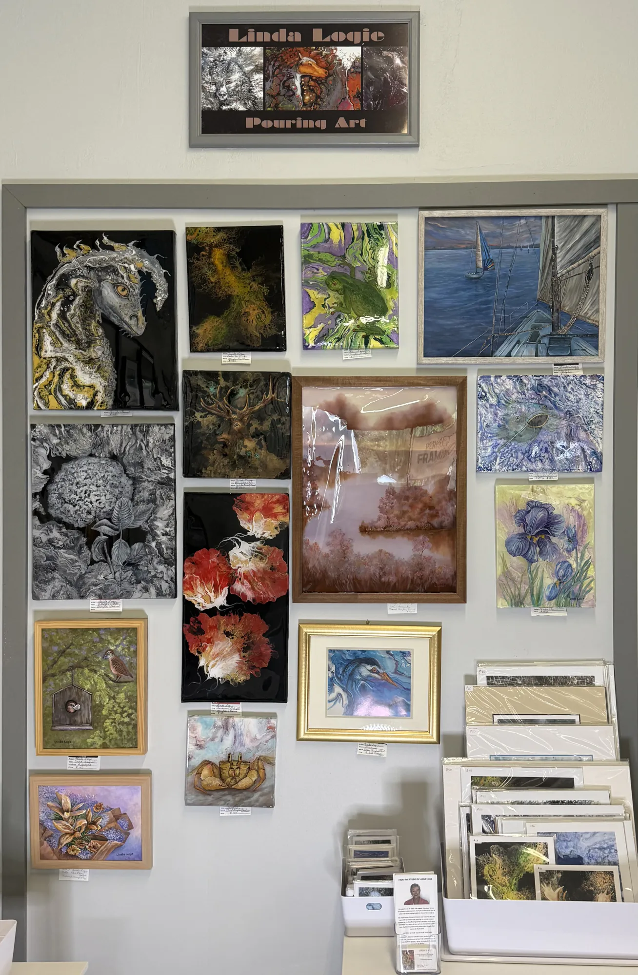 A display of various artworks including animals, nature scenes, and abstract designs. Framed, unframed pieces, and prints are organized on a wall and a table below.
