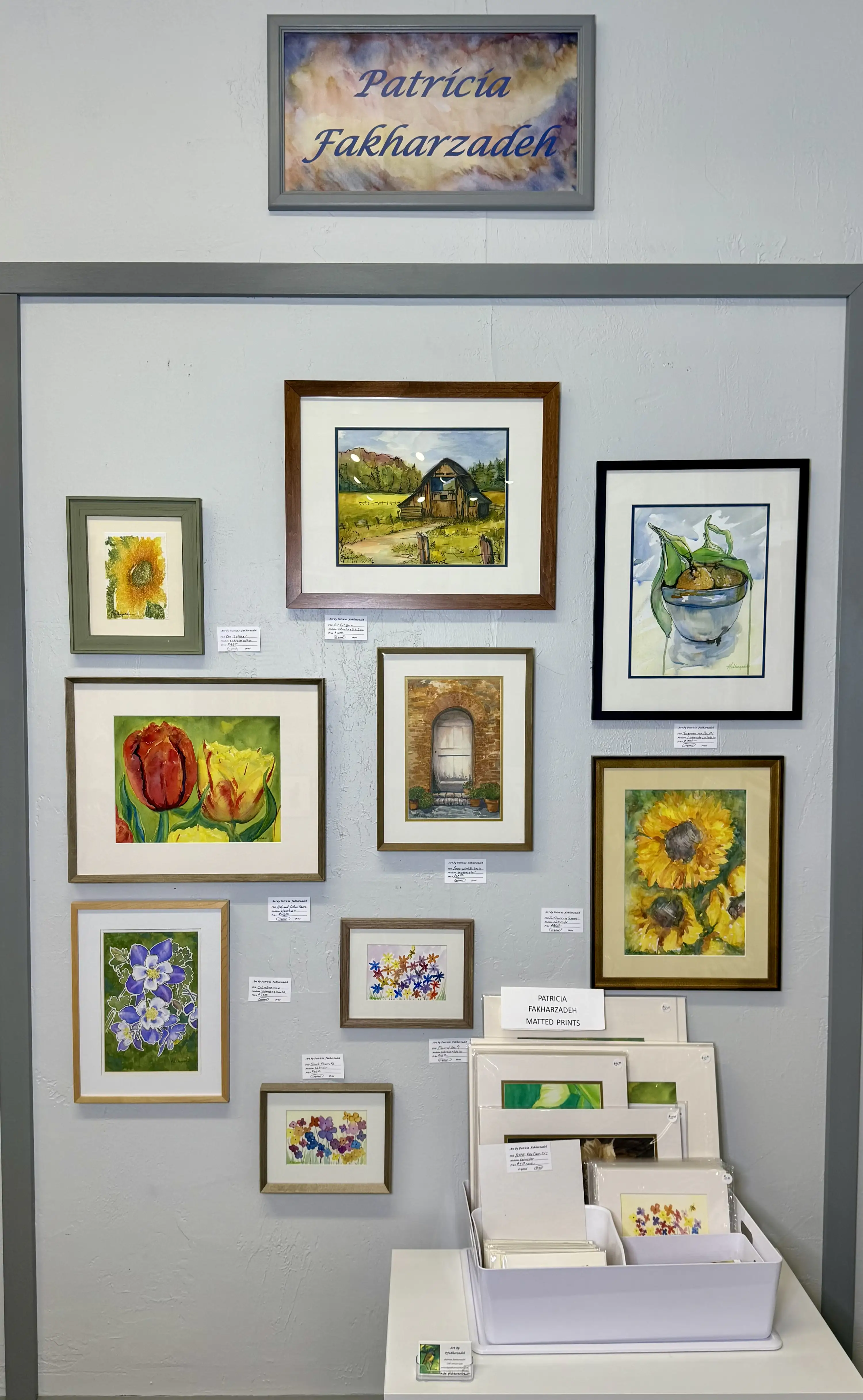 Wall display of framed watercolor artworks featuring flowers, nature scenes, and a building, with additional matted prints in a container below, labeled 