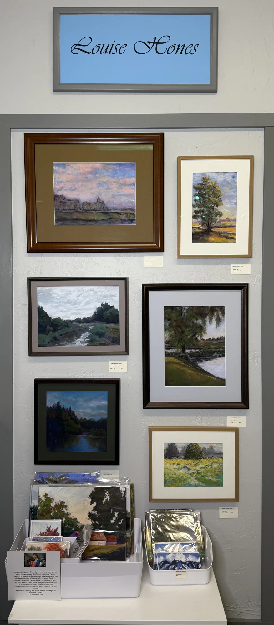 Art gallery wall featuring six framed landscape paintings by Louise Hones. Below, two bins hold smaller artworks and prints. A sign on top displays the artist's name.