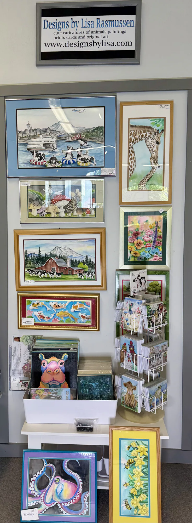 Wall display of colorful animal and nature artwork, including paintings of giraffes, cows, mushrooms, flowers, koi fish, an octopus, and bees. Greeting cards are on a rack below.