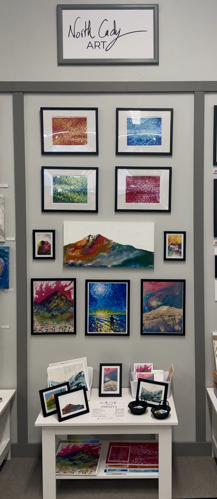 Gallery wall featuring colorful abstract paintings and prints in black frames. A small table below displays more framed art, prints, and a price list for 