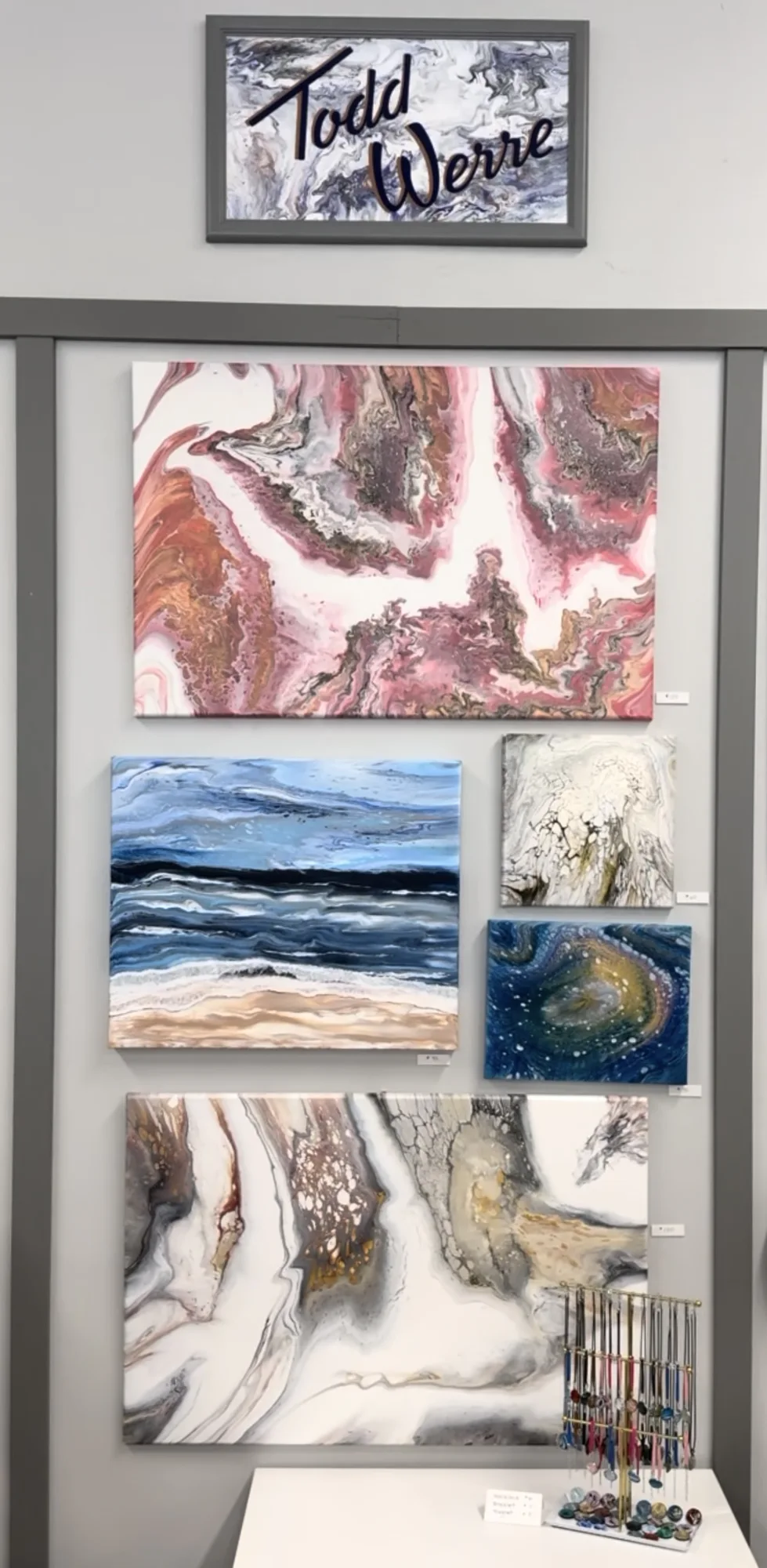 Art display featuring abstract paintings with swirls of pink, blue, and earth tones. A sign with the artist's name is above. Jewelry is displayed on a table below.