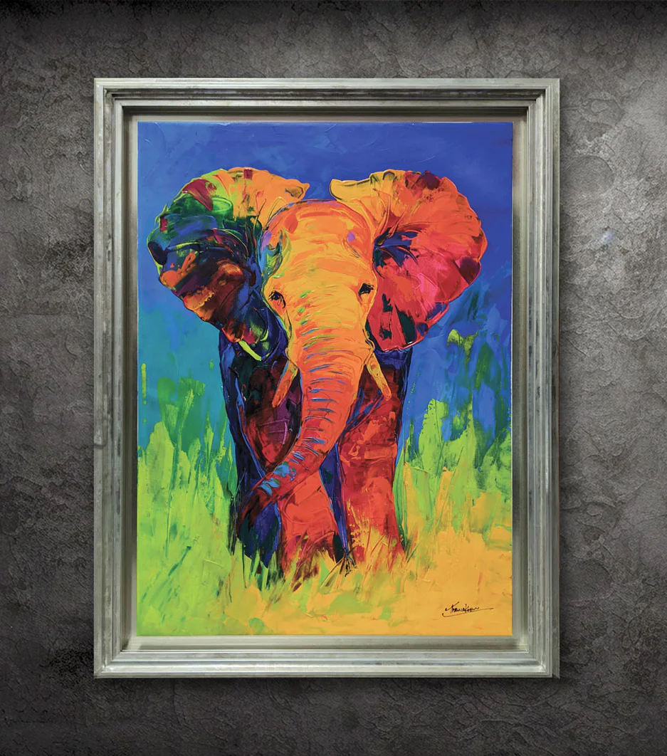 A colorful painting of an elephant with a multicolored trunk and ears, set against a green and blue background, framed in a silver frame, displayed on a textured gray wall.