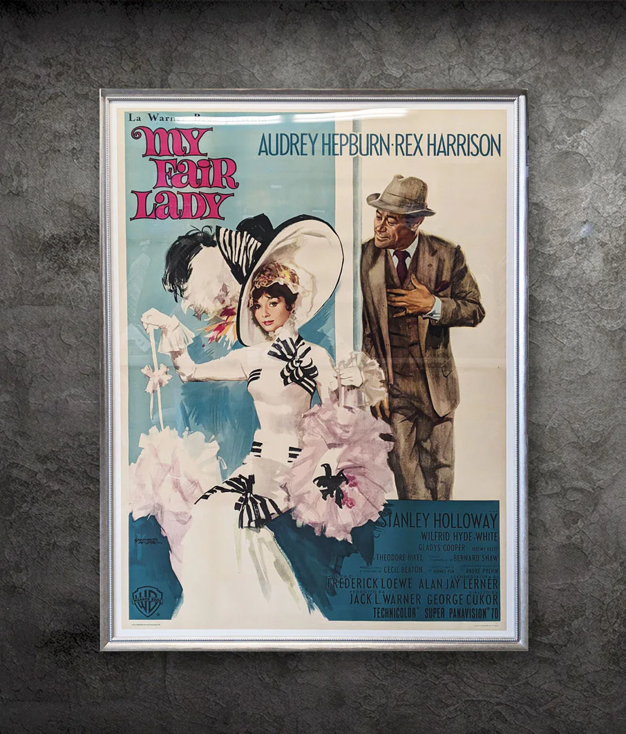 Framed poster of “My Fair Lady” featuring Audrey Hepburn in a white dress and large hat with ribbons, and Rex Harrison in a suit and fedora, against a textured gray wall.