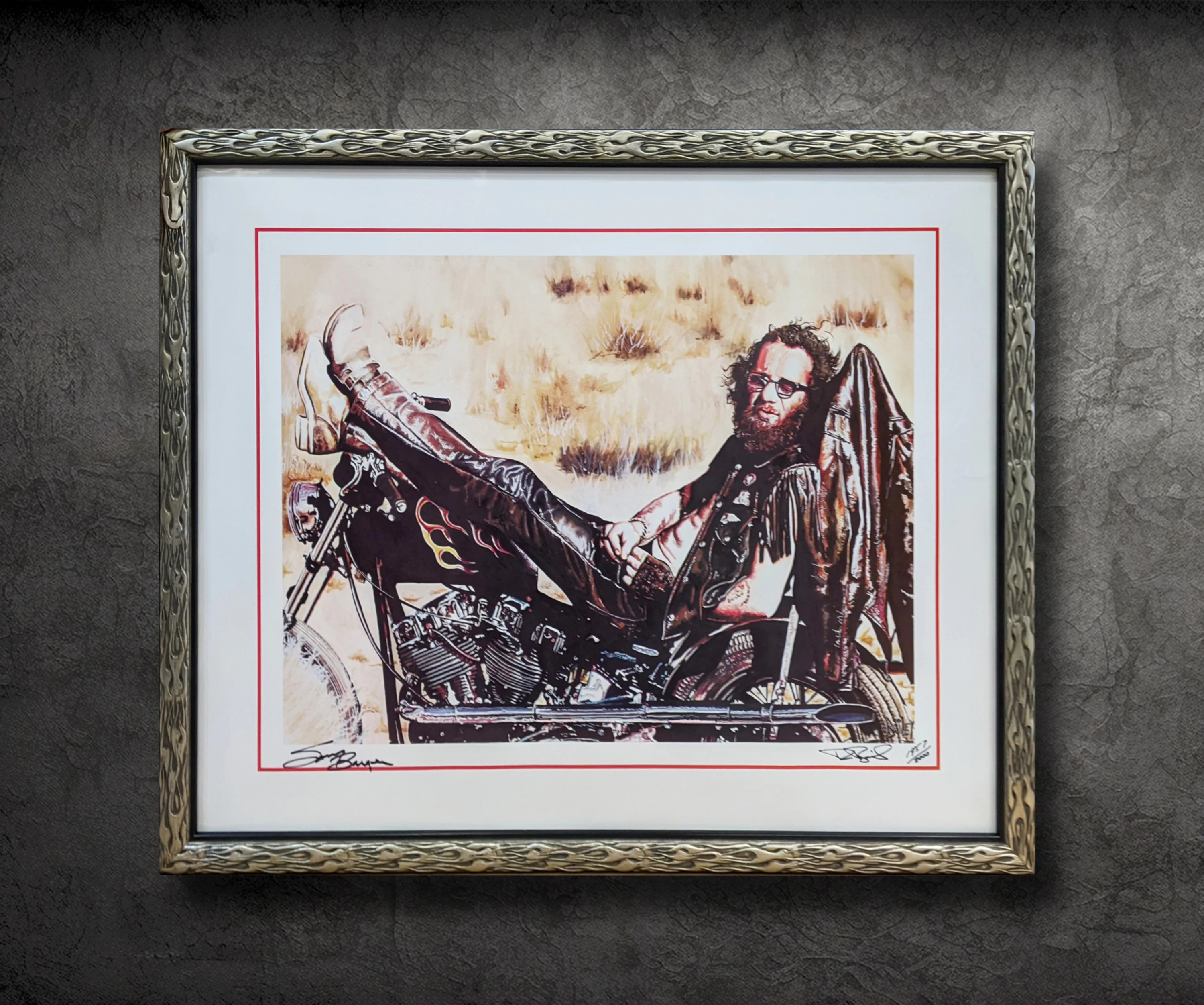 Framed artwork of a man with a beard and glasses lounging on a motorcycle in a desert setting, wearing leather attire and boots. The frame has an ornate design with a white mat border.