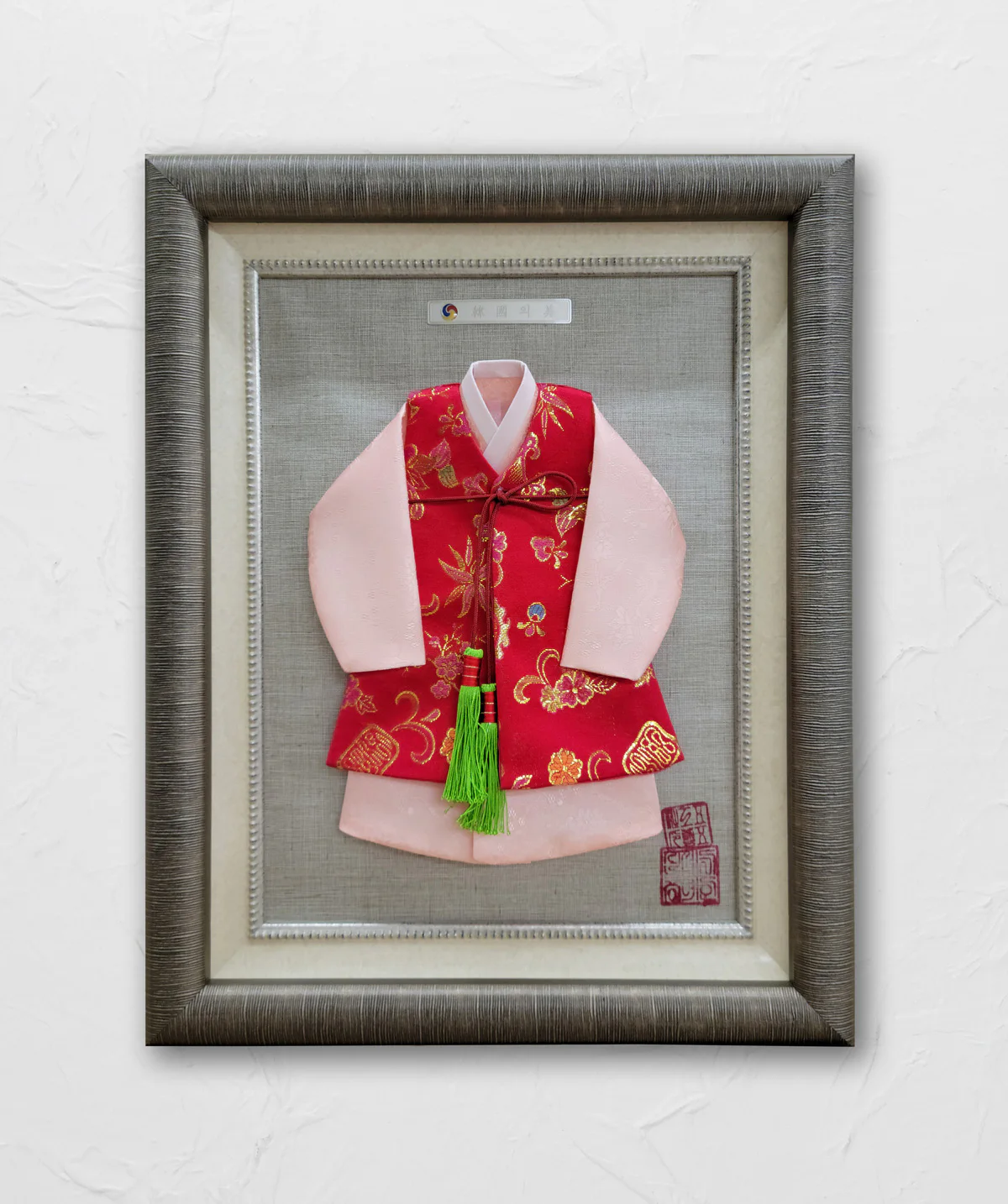 Framed traditional Korean child's hanbok displayed on a grey textured background. The outfit includes a red floral patterned vest over a pink robe and is adorned with a green tassel.