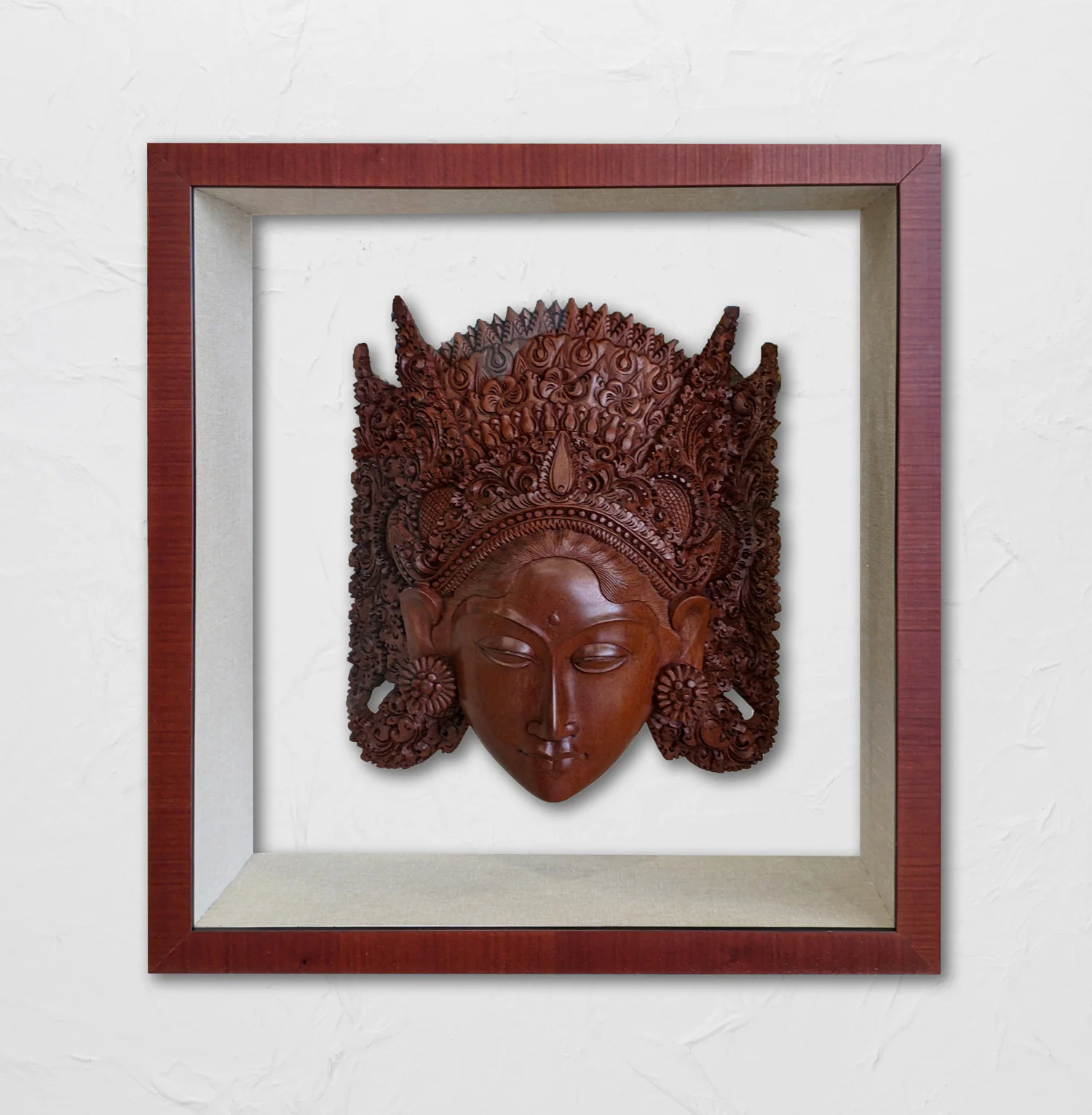 Wooden carved face mask with intricate headpiece details mounted in a shadow box frame with a red-brown outer border and light gray inner frame, hung on a white wall.