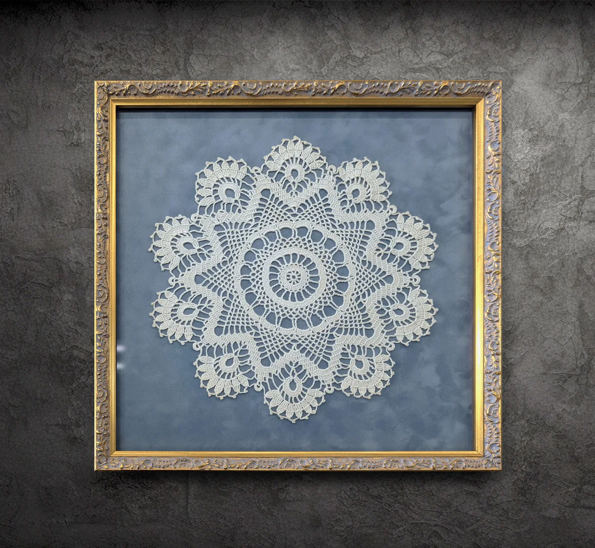 Framed white crochet doily with intricate patterns displayed against a blue background, hung on a textured grey wall. The frame is ornate and gold-colored.