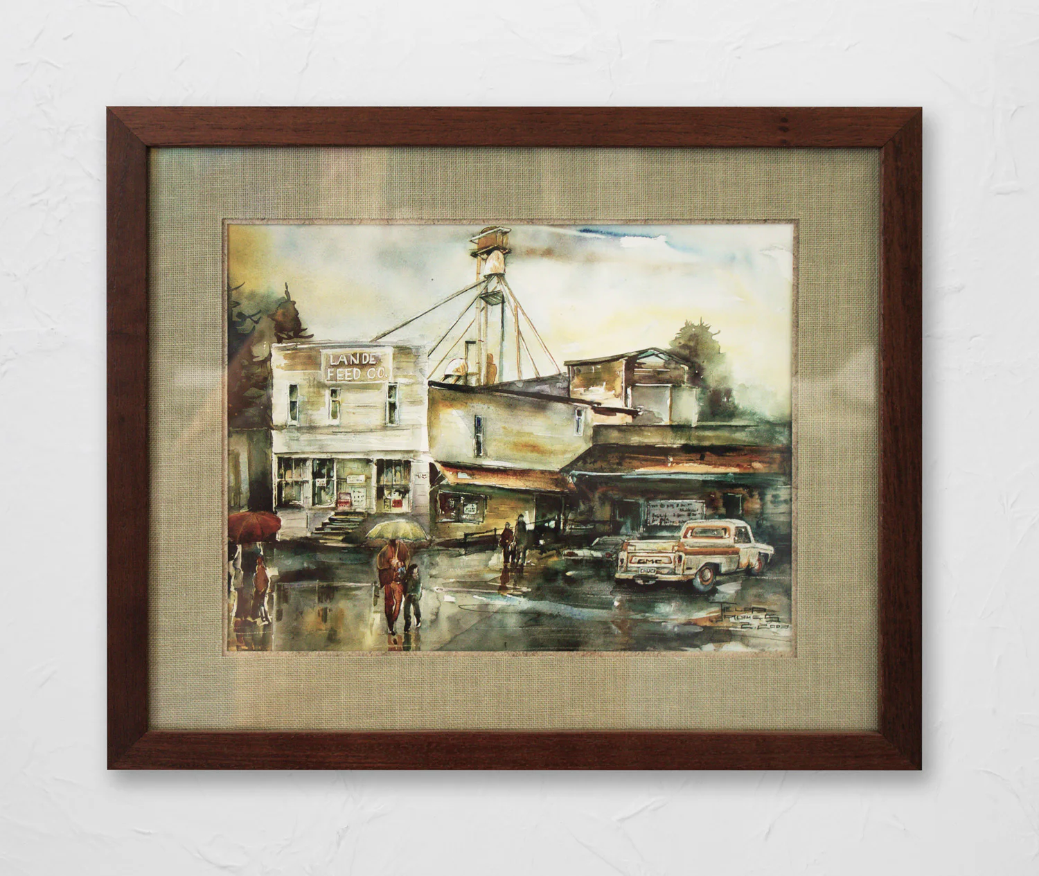Framed artwork depicting a wet street scene in front of a feed company, with people holding umbrellas, an old truck parked nearby, and industrial buildings in the background.
