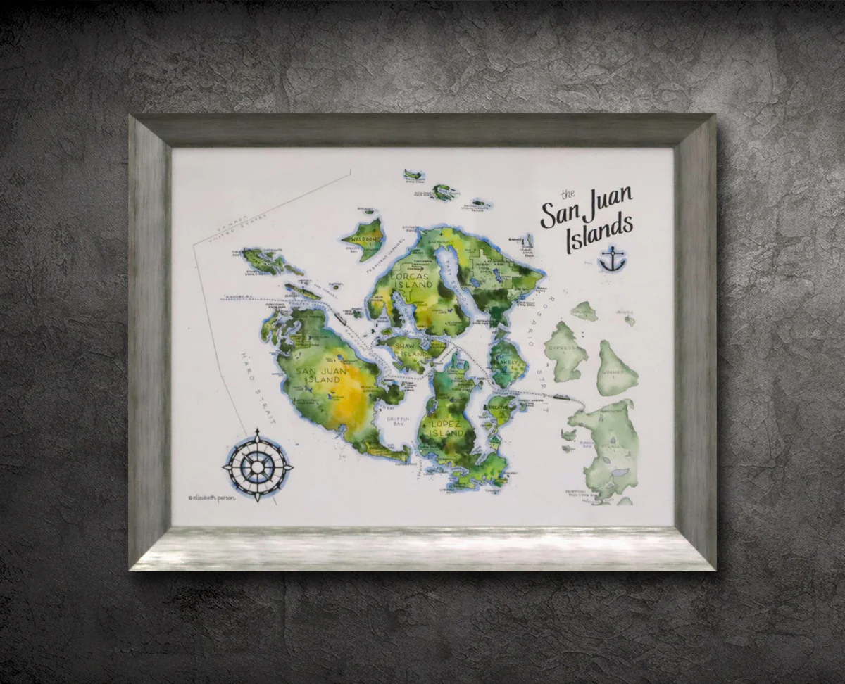 Framed watercolor map of the San Juan Islands on a textured gray wall. The map includes islands such as Orcas, San Juan, and Lopez, with a compass at the lower left corner.