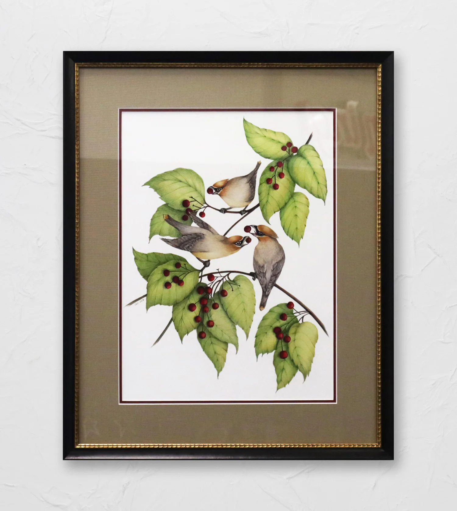 Framed artwork depicting three birds perched on a branch with green leaves and red berries. The frame is black with a golden inner edge, and the background is a textured white wall.
