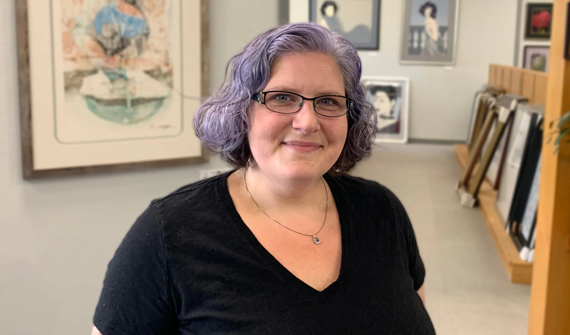 A person with purple hair and glasses is smiling, wearing a black top. They stand in an art gallery with various framed artworks on the walls and shelves in the background.