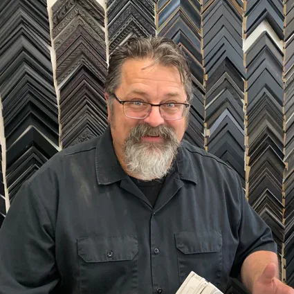 A person with glasses and a beard stands behind a counter displaying various picture frame corners. He is holding an open frame catalog, and there is a wall of frame samples in the background.