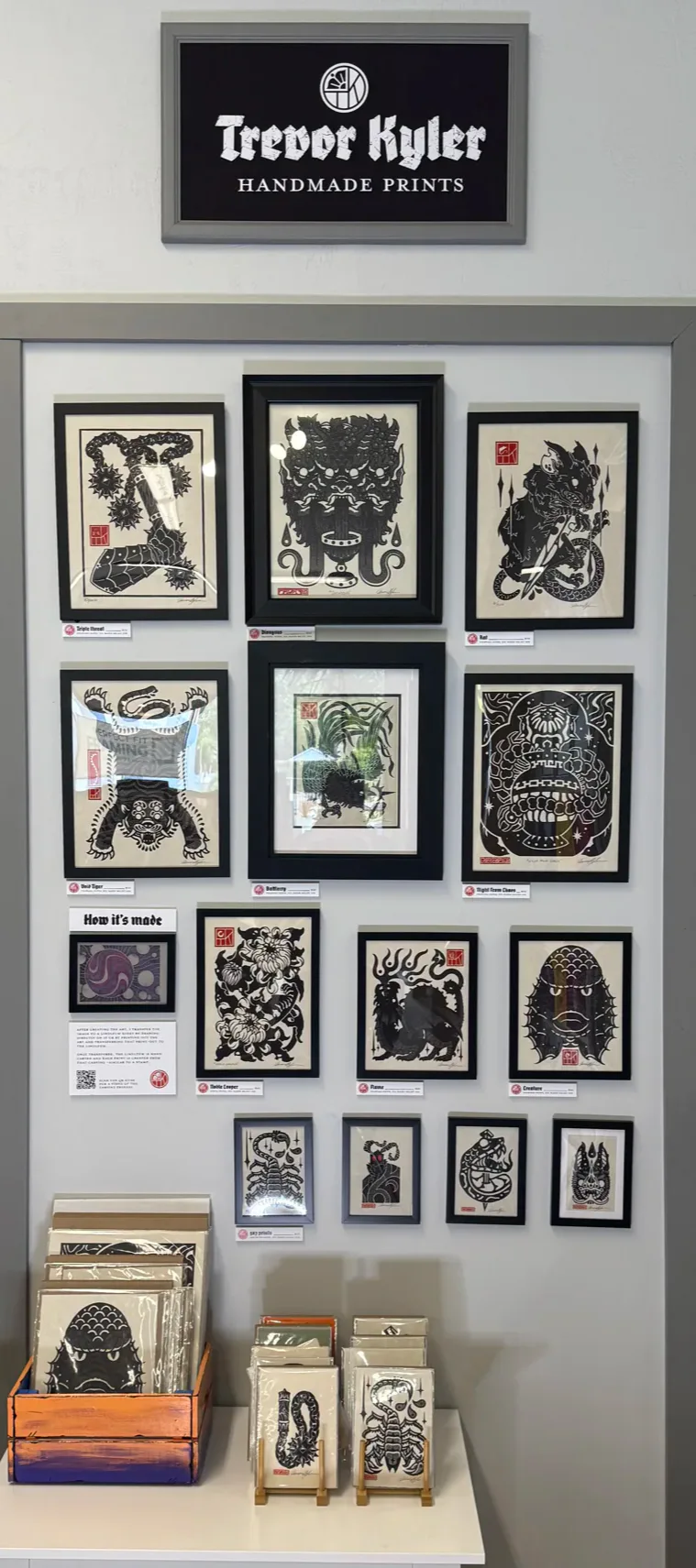 Display of Trevor Kyler's handmade prints on a wall with framed artwork in bold black-and-white designs. A table below holds smaller print items in various packaging.