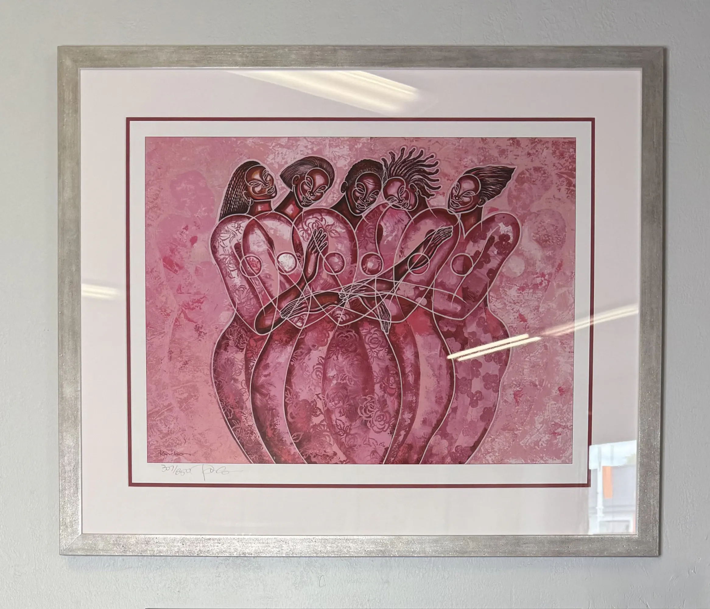 Framed artwork featuring abstract figures in pink and red hues, intertwined and facing forward. The background has a textured, floral pattern.