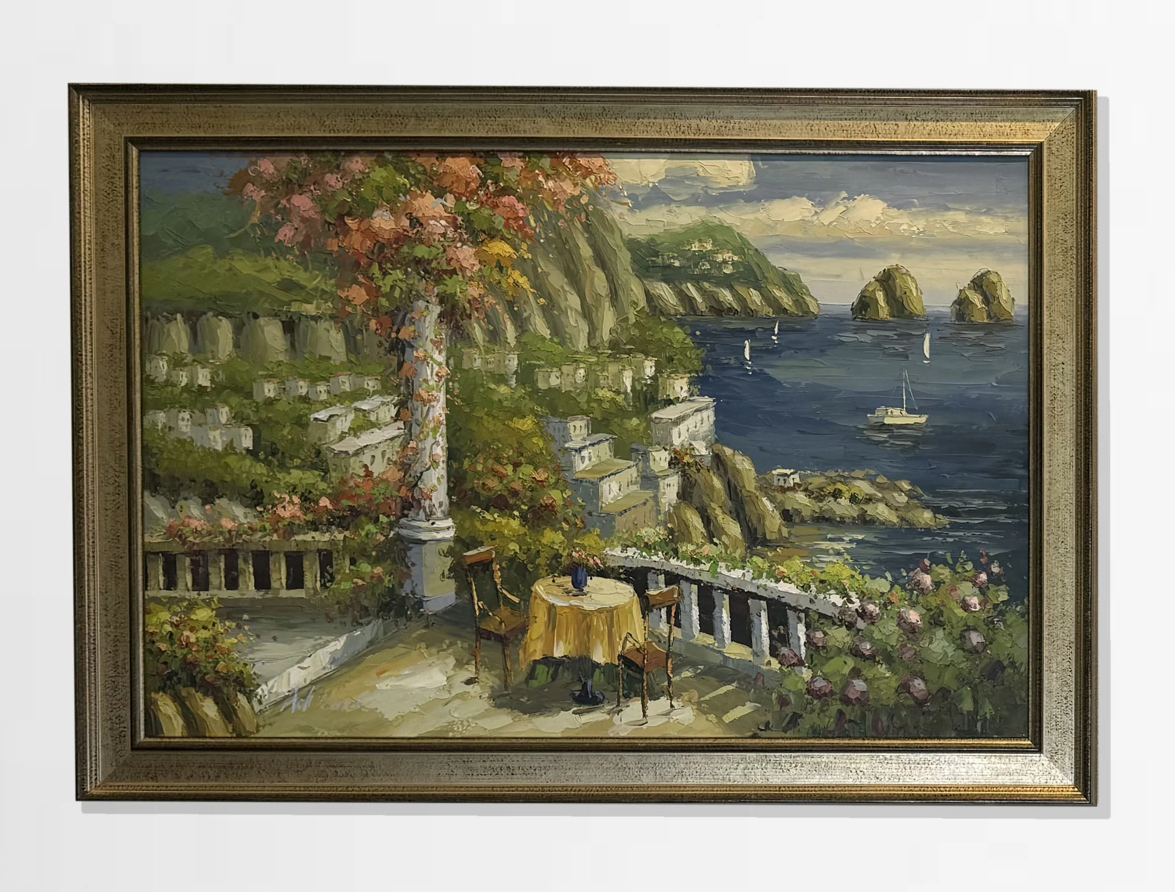 A framed painting depicts a scenic coastal view with cliffs, blue sea, sailboats, and distant islands. In the foreground, there are flowers, a column, and a table with two chairs.