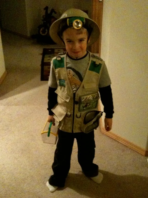 A child wearing a safari outfit stands in a hallway. The attire includes a safari hat with a light, vest with pockets, black sleeves, and pants, holding a small box.