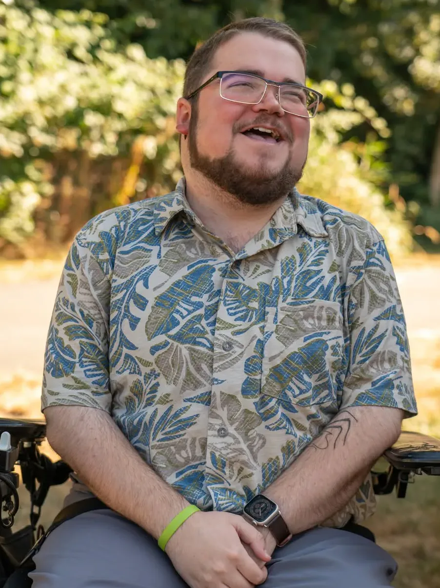A person with glasses and a beard smiles while sitting in a wheelchair. They wear a tropical-patterned shirt and a watch, with greenery in the background.