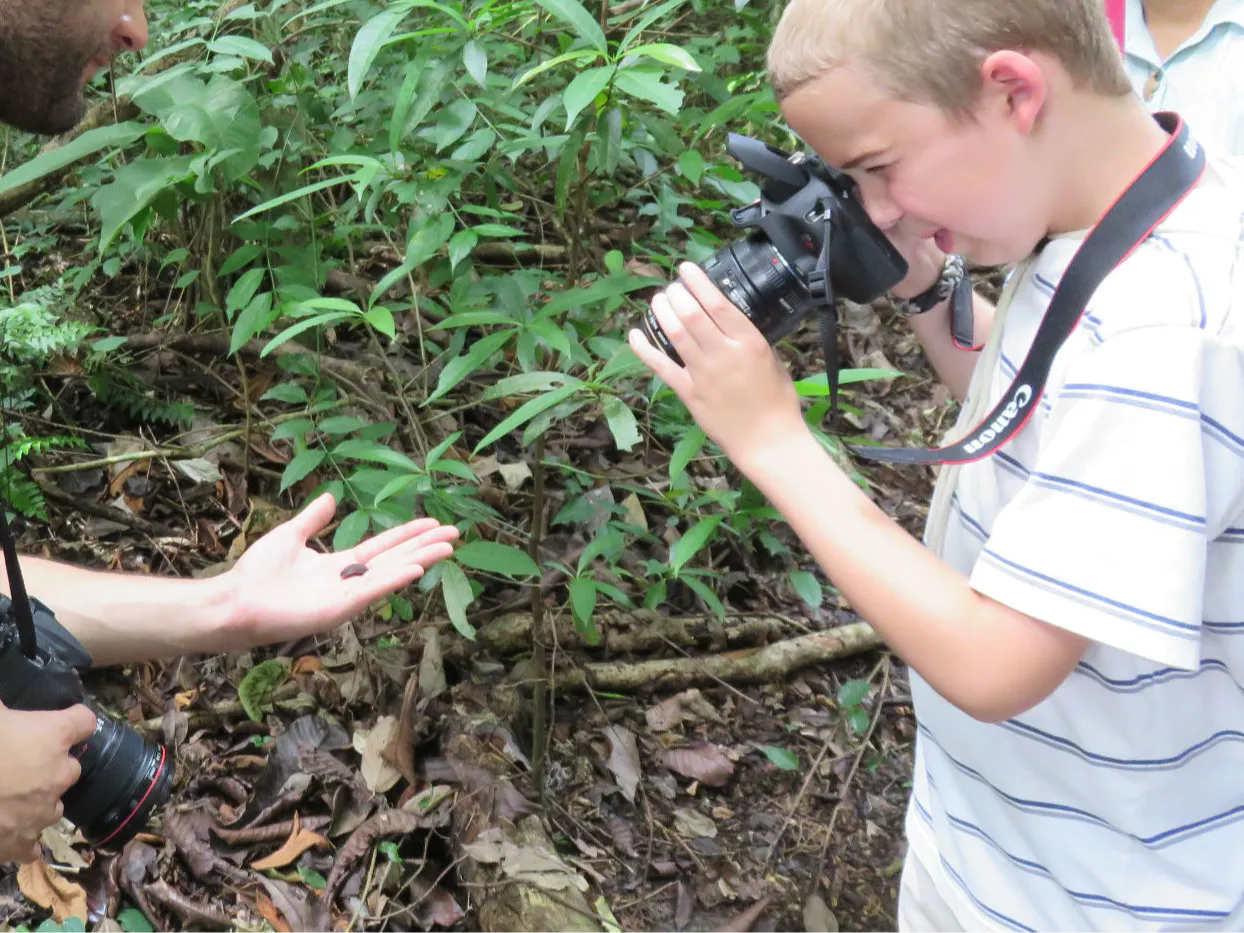 Boy photographing a beetle in a man's hand against a backdrop of green leaves and forest floor. Both hold cameras, focused on capturing the small insect in nature.
