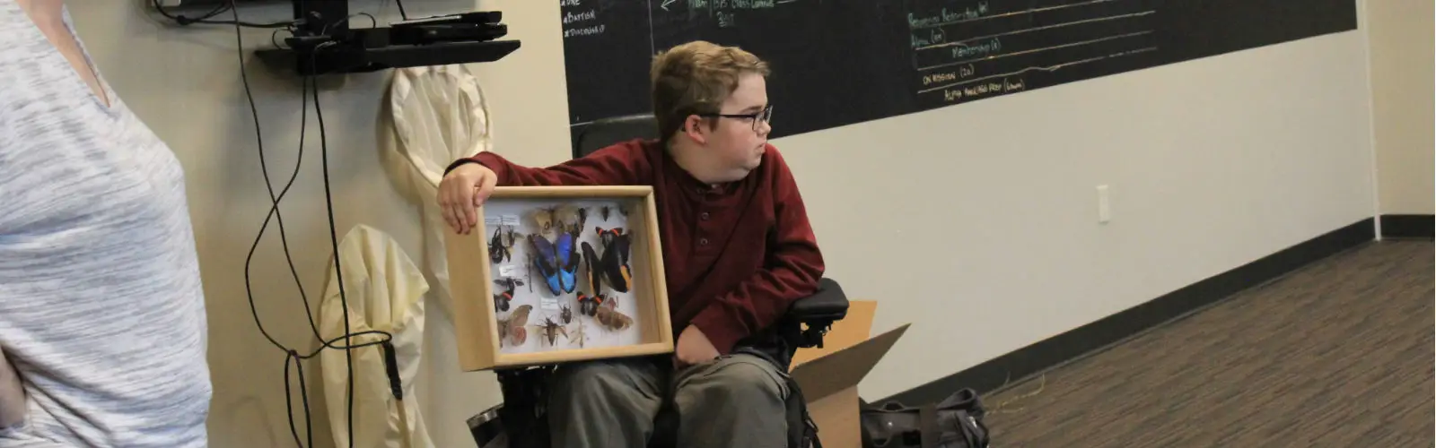 A person in a wheelchair holds a framed collection of butterflies and insects. A chalkboard with writing is in the background. Another person stands partially visible to the left.