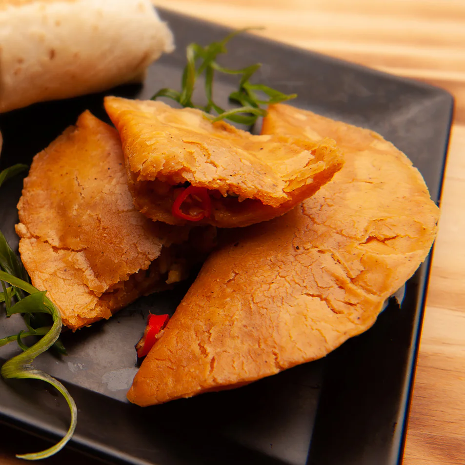 Golden empanadas filled with red peppers and vegetables served on a black plate, garnished with fresh green herbs, on a wooden table background.