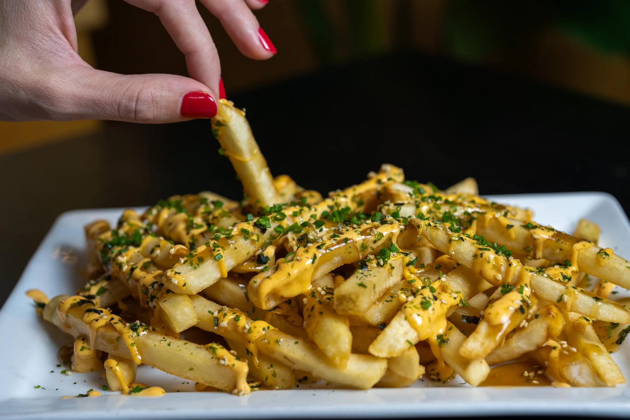 A hand with red nail polish picks up a seasoned french fry from a plate of fries drizzled with orange sauce and topped with chopped herbs, on a dark background.