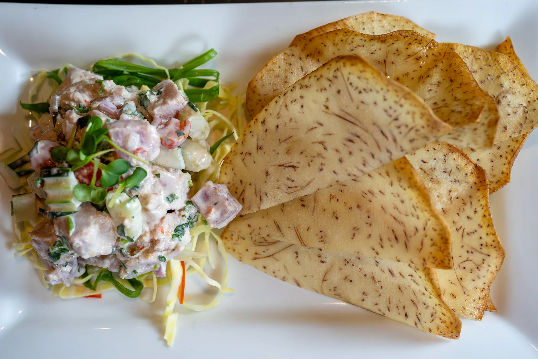 A dish with a creamy fish salad, including diced fish, cucumber, and greens, is served on a bed of cabbage next to slices of lightly browned taro chips on a white plate.
