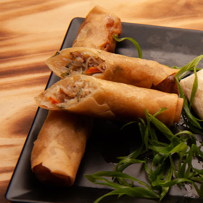 Three crispy spring rolls on a black plate with sliced green onions, one cut in half to show the filling of noodles and vegetables, set on a wooden table.