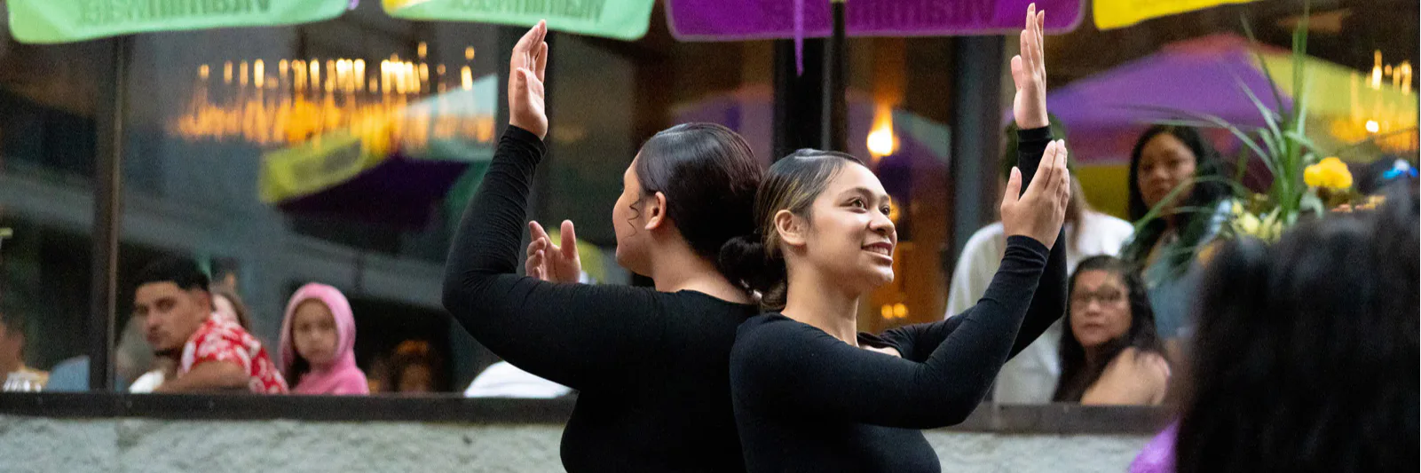 Two people in black outfits perform a synchronized dance outdoors, with colorful umbrellas and string lights above. A small crowd watches in the background.