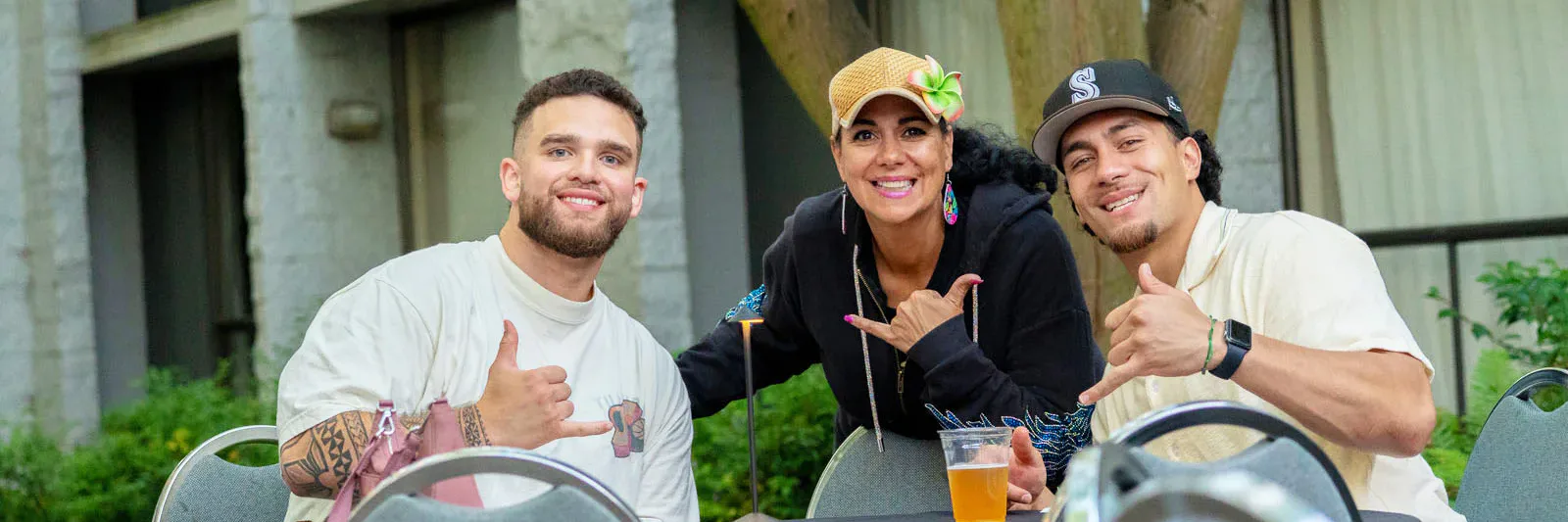 Three people sitting at an outdoor table, smiling and making a shaka hand gesture. They are in a garden setting with trees and a building in the background.