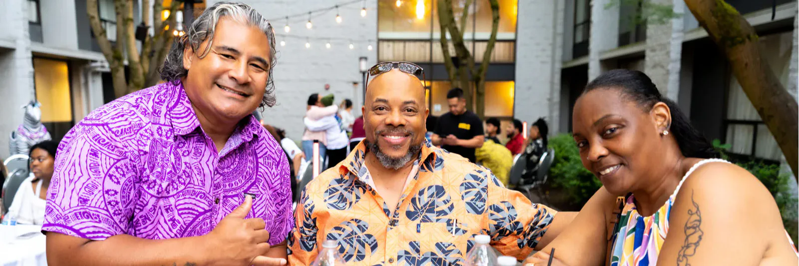 Three people smiling at a table outdoors, wearing colorful shirts. The table has drinks and a green leaf decoration. String lights and trees are visible in the background.