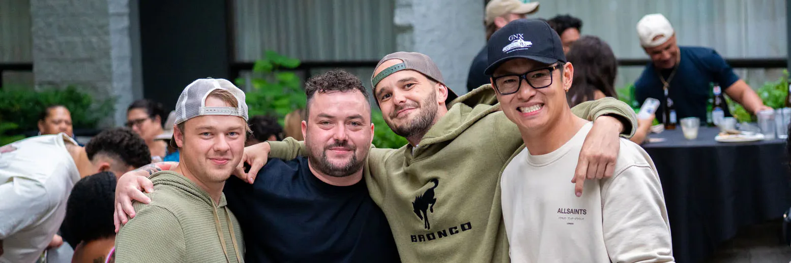 Four men smiling and posing together outdoors at a gathering. They are dressed casually, with others socializing and a table with drinks in the background.