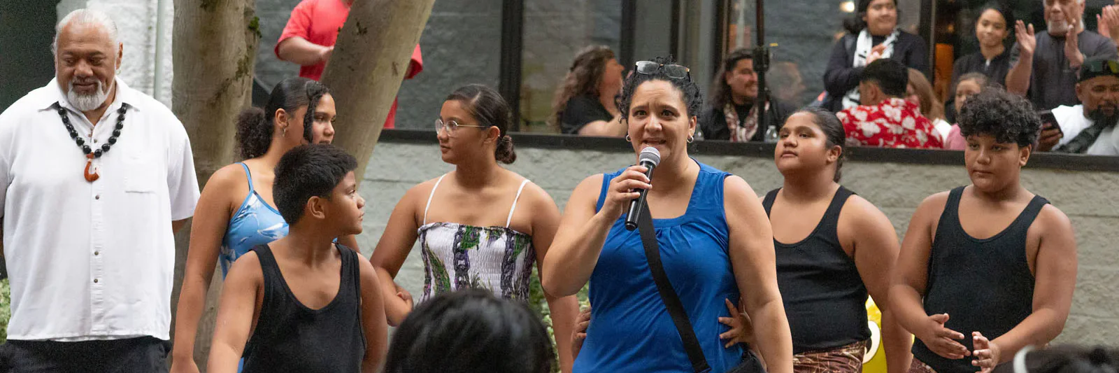 A group of people in traditional attire performs outdoors; a woman in a blue dress holds a microphone, while others sing and dance, with onlookers in the background.