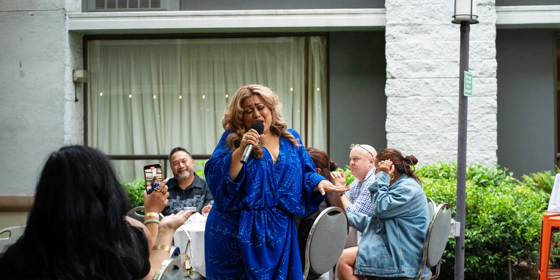 A performer in a blue dress sings into a microphone at an outdoor event. Audience members sit at tables, with one person taking a photo on their phone.