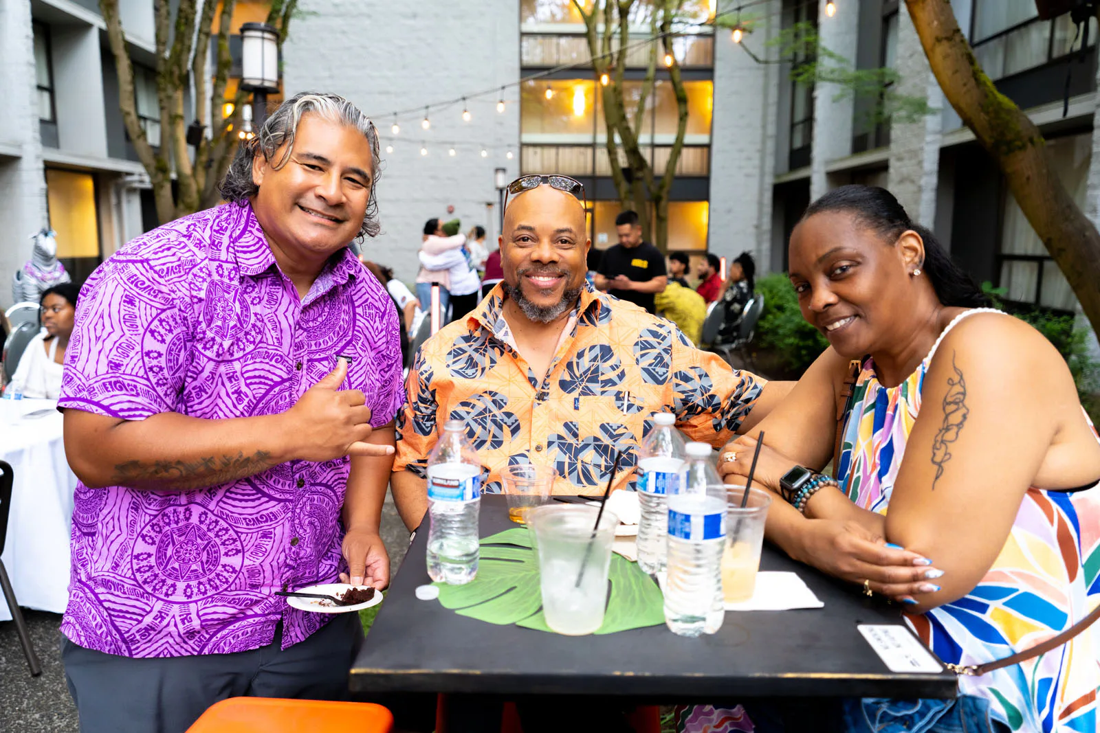 Three people smiling at a table outdoors, wearing colorful shirts. The table has drinks and a green leaf decoration. String lights and trees are visible in the background.