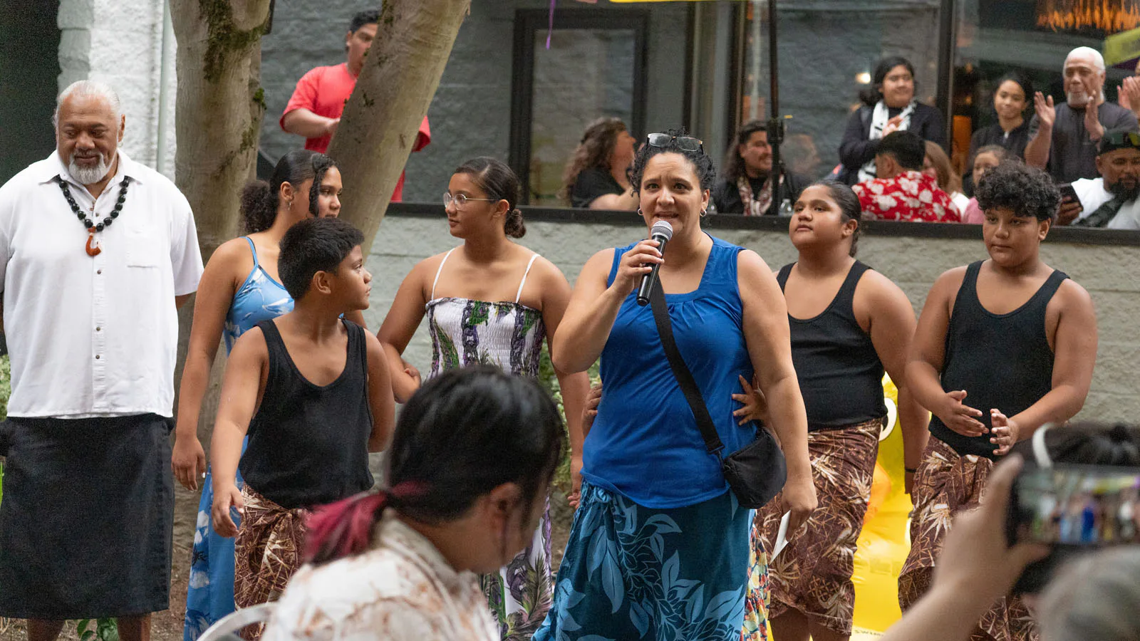 A group of people in traditional attire performs outdoors; a woman in a blue dress holds a microphone, while others sing and dance, with onlookers in the background.