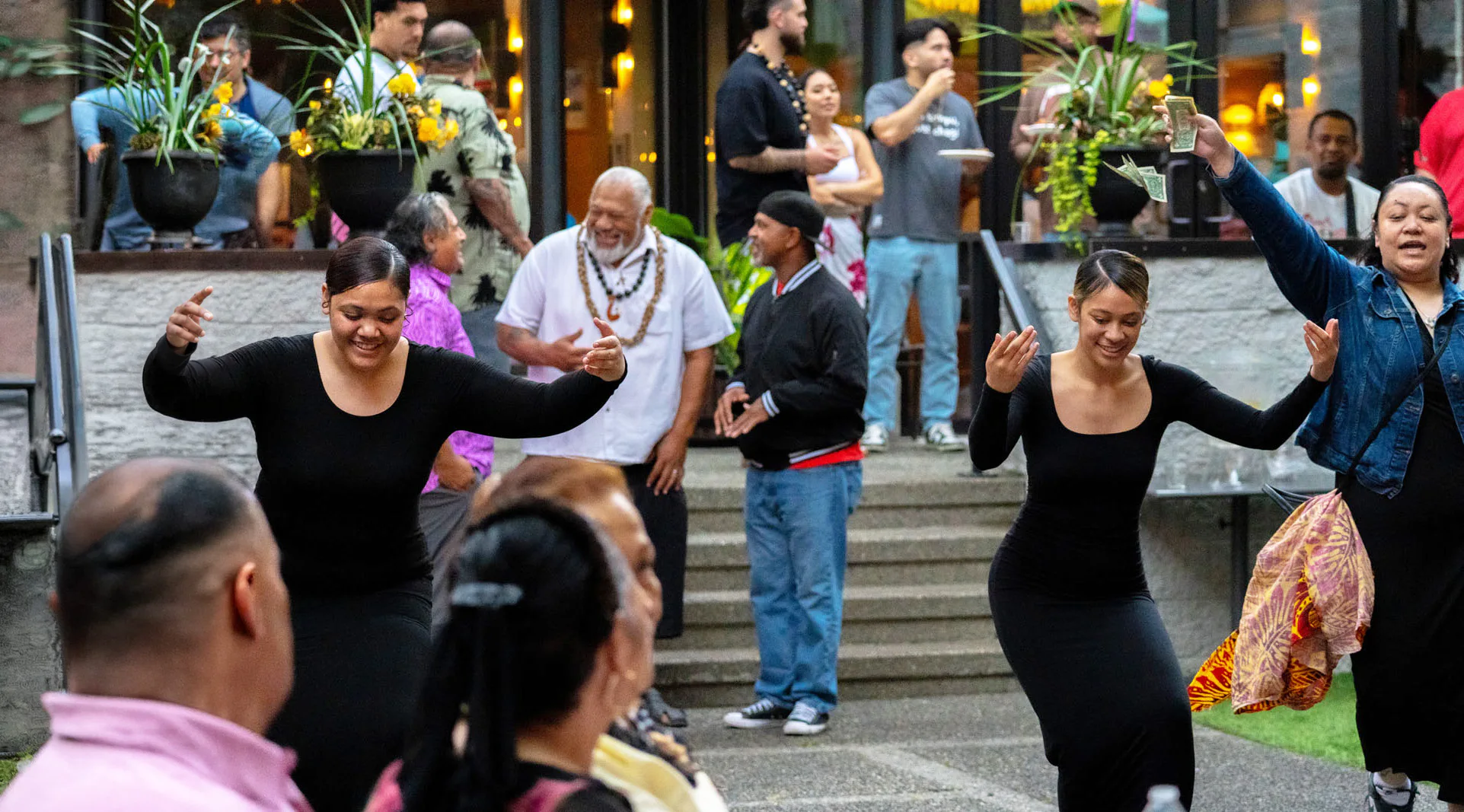 Two women in black dresses dance joyfully in an outdoor gathering. A woman in a denim jacket holds up money. People in the background watch and smile near potted plants and stairs.